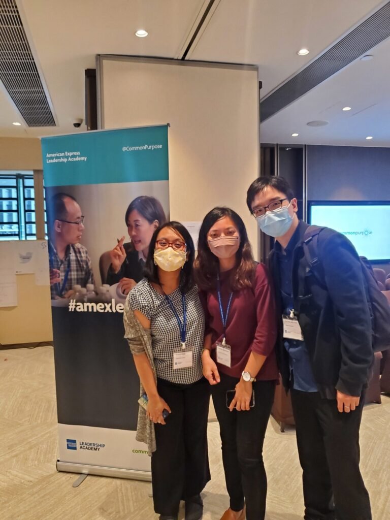 Three participants of the American Express Leadership Academy posing together in an indoor event space, all wearing face masks and name tags. A promotional banner for the Leadership Academy and Common Purpose is visible in the background.