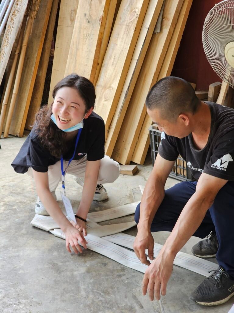 A woman with a big smile kneeling on the floor, wearing a black shirt and a blue lanyard, alongside a man assisting her with wide strips of fabric. The setting includes wooden planks and a fan, suggesting a workshop or community building environment.