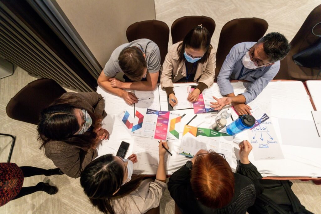 A top-down view of a group of six individuals collaborating at a table covered with colorful brochures, handwritten notes, and water bottles. Participants wear masks and are actively engaged in discussion.