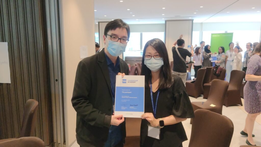 Two individuals, both wearing face masks, holding a certificate of participation from the American Express Leadership Academy in an indoor event setting. They are standing close together, smiling behind their masks, with chairs and participants visible in the background.